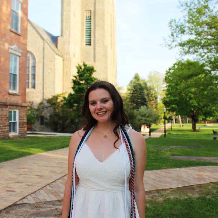 Young White woman with brown hair standing in front of the chapel at St. Lawrence University, smiling and posing in her white graduation dress with honor cords around her shoulders.