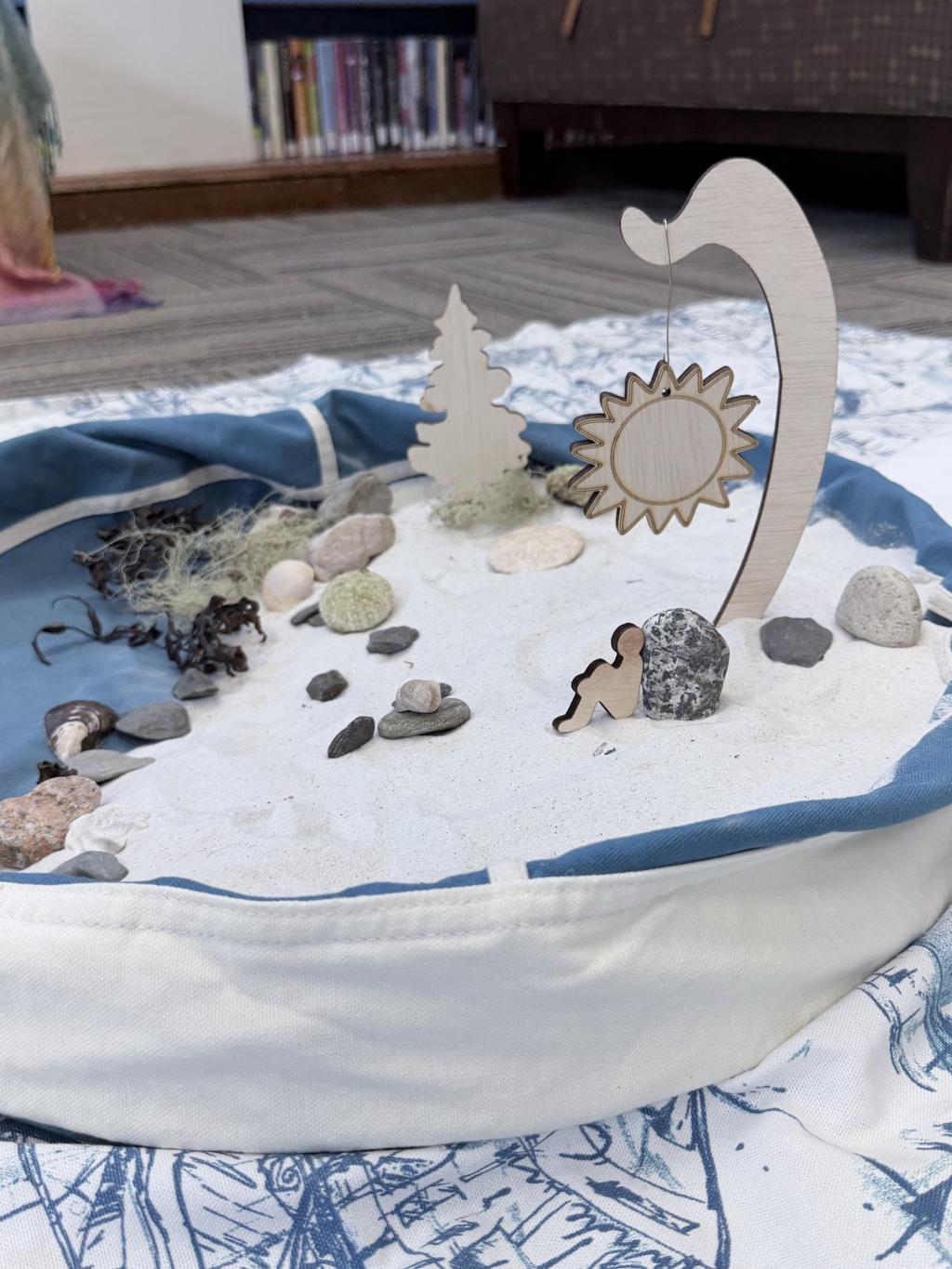 Rocks and wooden figurines arranged in a small blue vessel with sand to replicate a beach scene.