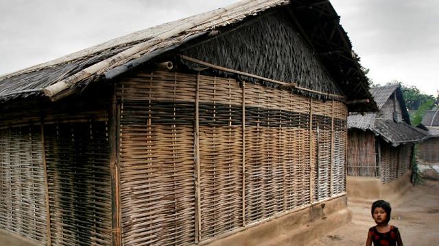 It is the picture of a bamboo hut in the Bhutanese refugee camp in Jhapa, Nepal. A young girl is standing at the corner of the hut while looking at the camera. The sky is full of monsoon clouds.