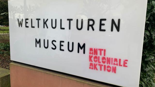 A white sign with black text and red graffiti in front of a museum.