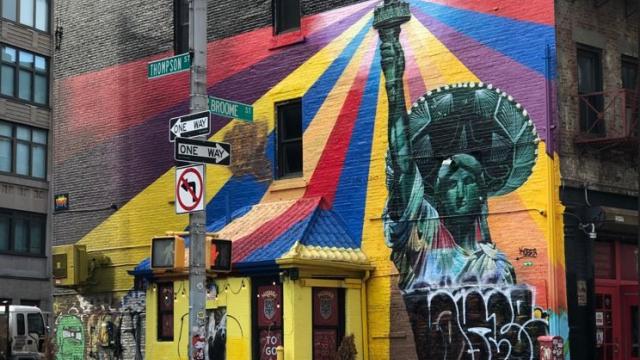 Street art depicting the statue of liberty wearing a sombrero with rainbows emanating from her torch.