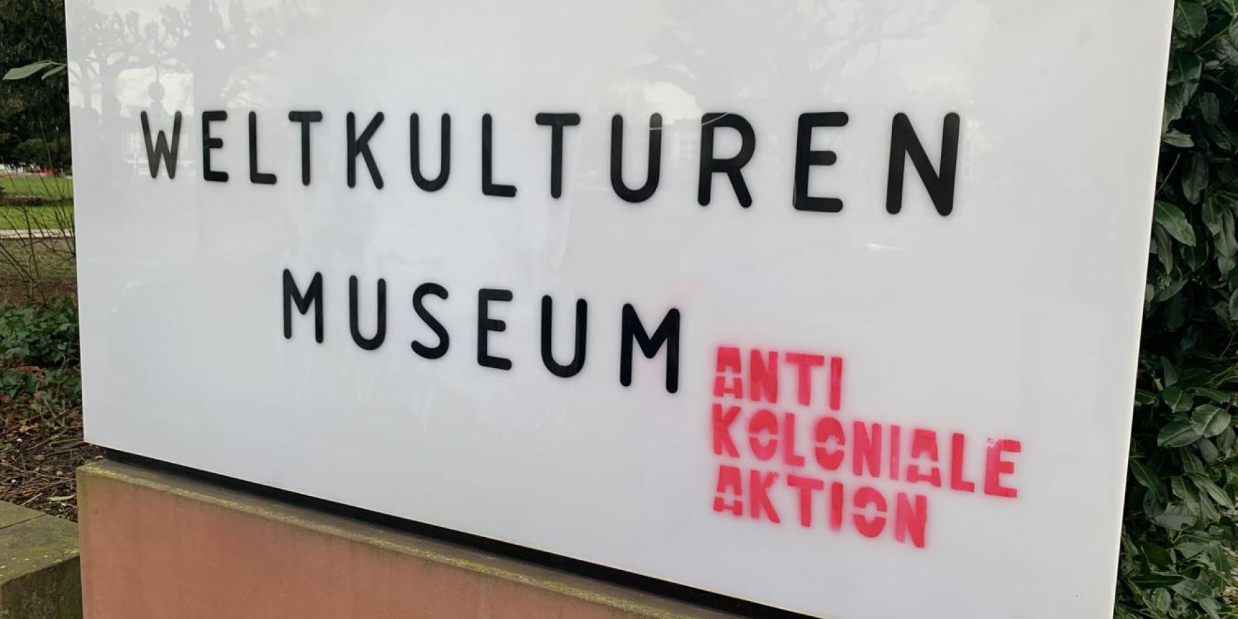 A white sign with black text and red graffiti in front of a museum.
