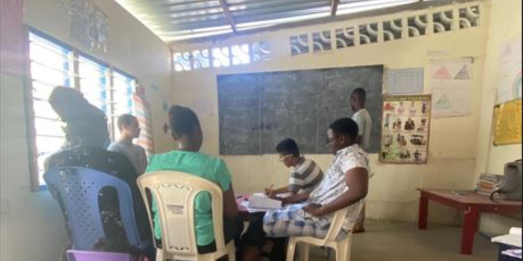 Four people sit at a table while another person stands by a chalkboard.