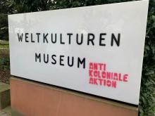 A white sign with black text and red graffiti in front of a museum.