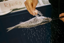 A hand sprinkling salt onto a fish on a stick.