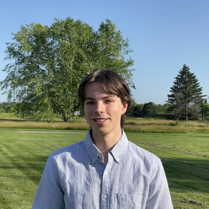 A picture of Alex from the waist up. He is outside and there are grass, trees, and blue sky in the background. He is smiling.
