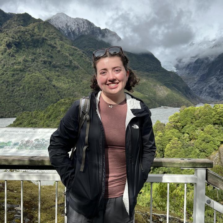 Woman with brown hair wearing a raincoat and hiking cloths standing in front of mountains.