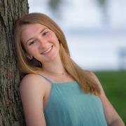 Haylei has long, blonde hair and blue eyes. She is seen here leaning against a tree and smiling.
