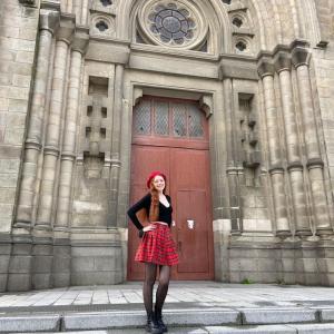 Alexandra stands in a black shirt and plaid skirt in front of an ornate stone door.