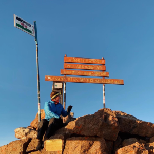 Anna sits at the top of Mount Kenya next to a sign.