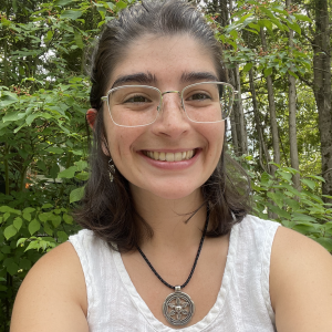 Image of smiling Ania. She has shoulder-length black hair tied in a half ponytail. She wears square silver glasses, silver earrings, and a necklace made of black thread with a flower-shaped pendant. She is also wearing a white linen top. In the background, we can see green vegetation, mostly leaves and some tree trunks. 