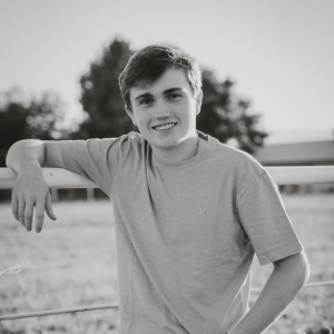 A photo of Blake leaning up against a fence with a field and trees behind him. 
