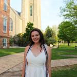 Young White woman with brown hair standing in front of the chapel at St. Lawrence University, smiling and posing in her white graduation dress with honor cords around her shoulders. 