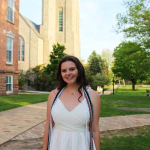 Young White woman with brown hair standing in front of the chapel at St. Lawrence University, smiling and posing in her white graduation dress with honor cords around her shoulders. 